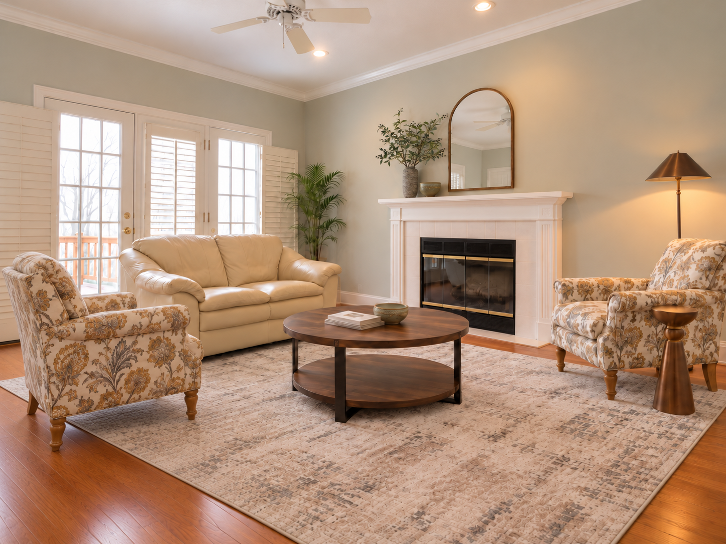 The same living room redesigned around the cream leather loveseat, with two patterned armchairs, a round wood coffee table, a large neutral rug, an arched mirror over the fireplace and a brass floor lamp.