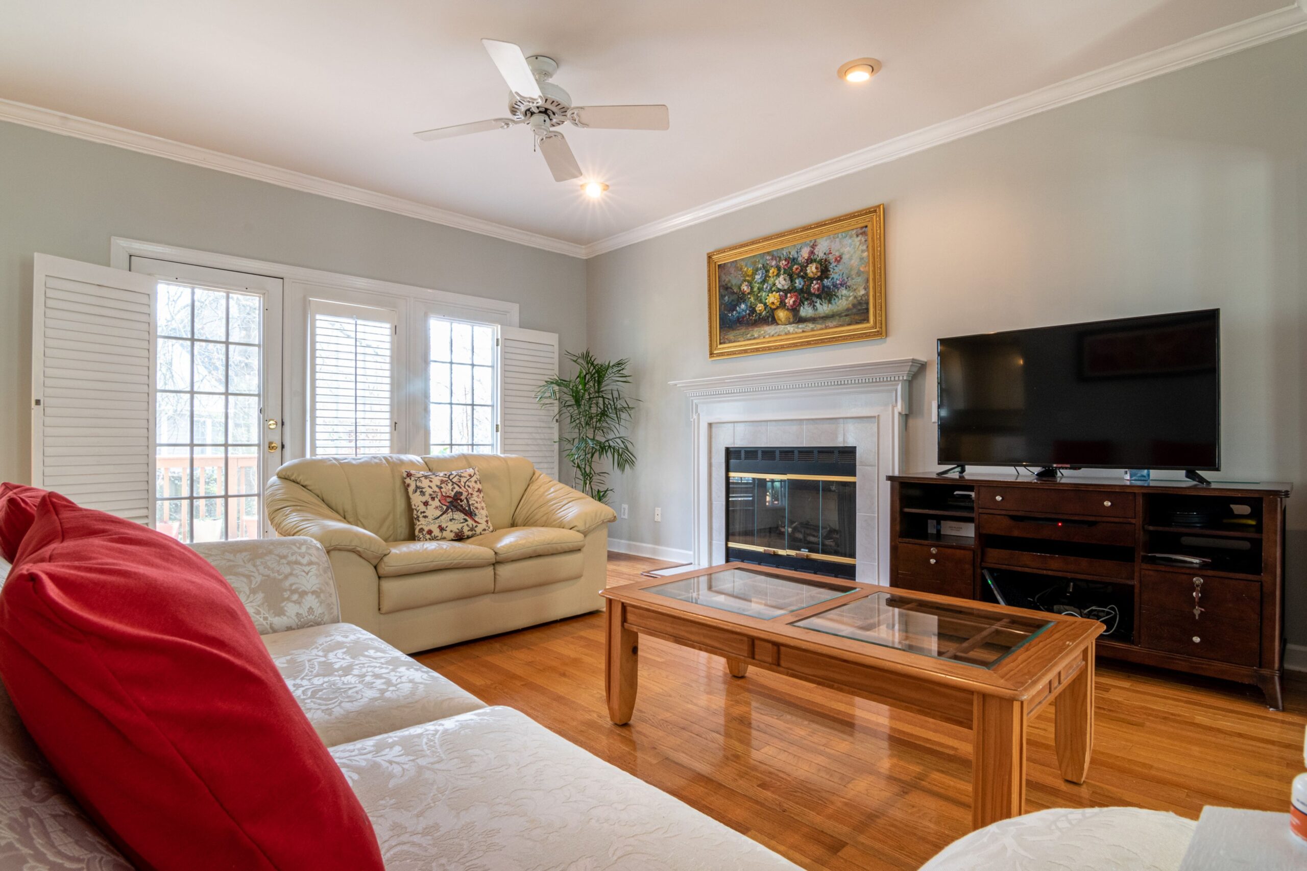 Living room with hardwood floors and a white fireplace, furnished with a cream leather loveseat, a damask sofa, a glass-top coffee table and a TV console beside the fireplace, with no rug.